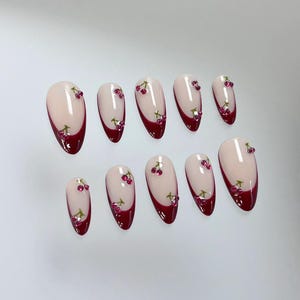 May include: Set of ten false nails with a cherry design. The nails are oval shaped and have a white base with a burgundy tip. Each nail has a small cherry design with green leaves and pink rhinestones.