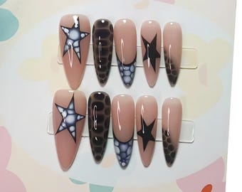 May include: A set of ten almond-shaped false nails. The nails feature a nude base with various designs, including a white and navy blue star, a dark brown snakeskin pattern, and a black star. Some nails have a dark gradient.