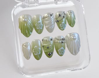 Op de afbeelding: Een set van tien groene en witte kunstnagels met parel- en zilveren versieringen in een doorzichtige plastic container.