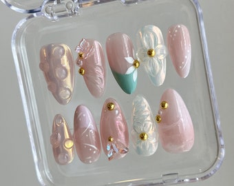 May include: A set of ten pink and white press-on nails with floral designs and gold accents. The nails are arranged in a clear plastic case.