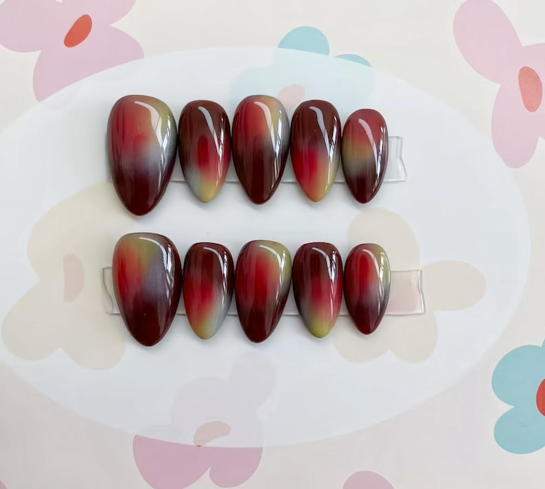 May include: A set of ten oval-shaped false nails with a gradient design. The colors transition from a deep burgundy at the base to a lighter yellow-green at the tips, with a touch of white in between. The nails are arranged in two rows on a white surface.
