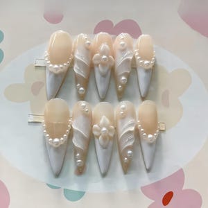 May include: Set of ten false nails with a pearl and swirl design. The nails are a light beige color with white swirls and pearl accents.