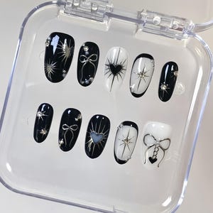 May include: A set of ten artificial nails in a clear plastic case. The nails feature black and white designs with silver accents, including bows, hearts, and starbursts. Some nails have small, sparkling rhinestones.