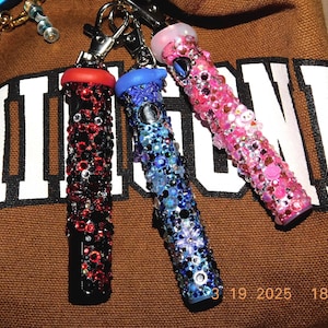 May include: Three cylindrical lighters decorated with colorful rhinestones and attached to silver metal swivel clasps. One lighter features red and black stones, one has blue and purple stones, and one is covered in various shades of pink stones.