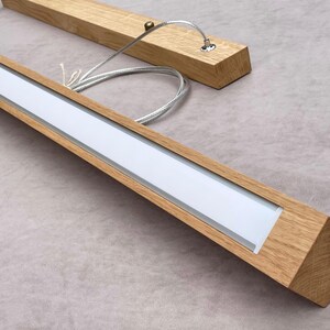 May include: Wooden light fixture with a rectangular design. The fixture has a light-colored wood frame with a white light panel. The fixture is suspended by thin metal cables.
