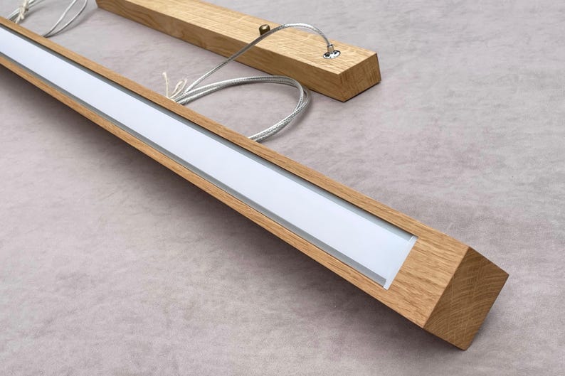 May include: Wooden light fixture with a rectangular design. The fixture has a light-colored wood frame with a white light panel. The fixture is suspended by thin metal cables.