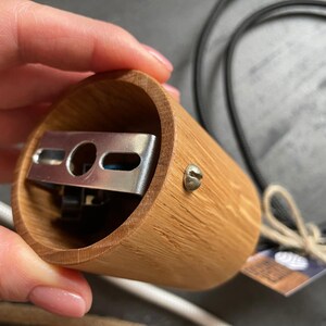 May include: A cylindrical wooden lamp holder with a metal bracket inside. The holder is made of light brown wood and has a screw on the side. A black cord and a white cord are visible in the background.