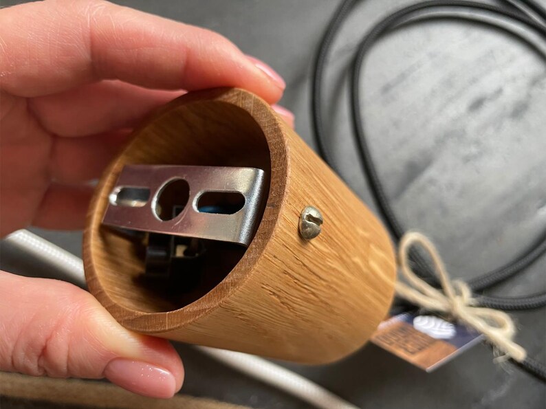 May include: A cylindrical wooden lamp holder with a metal bracket inside. The holder is made of light brown wood and has a screw on the side. A black cord and a white cord are visible in the background.