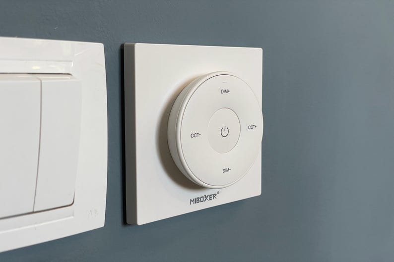 May include: A white wall-mounted light switch and a circular control panel. The control panel has a central power button and labels for dimming and color temperature adjustment. The brand name "MiBoxer" is printed on the panel.