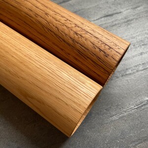 May include: Close-up of two rolled wood pieces. One is a light oak color, and the other is a darker, reddish-brown wood. The wood grain is visible on both pieces. The background is a gray, textured surface.