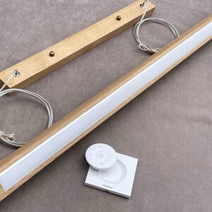 May include: A modern wooden LED light fixture with a white light strip. Includes a remote control and a square base. The fixture is made of light-colored wood and has a sleek design. The light is suspended by metal wires.