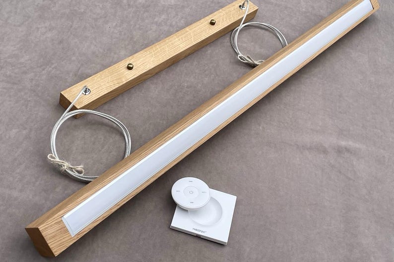 May include: A modern wooden LED light fixture with a white light strip. Includes a remote control and a square base. The fixture is made of light-colored wood and has a sleek design. The light is suspended by metal wires.