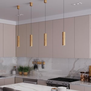 May include: A modern kitchen with light wood pendant lights hanging above a marble countertop island. The kitchen features light-colored cabinets, a stainless steel stove, and a sink with a black faucet. Shelves are stocked with books and decorative items.
