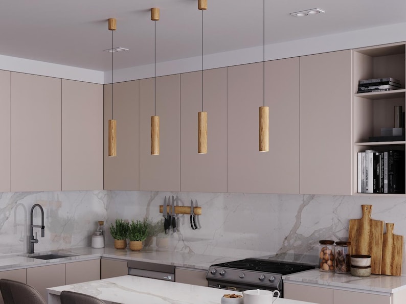 May include: A modern kitchen with light wood pendant lights hanging above a marble countertop island. The kitchen features light-colored cabinets, a stainless steel stove, and a sink with a black faucet. Shelves are stocked with books and decorative items.