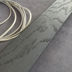May include: Close-up of a dark gray wooden plank with a textured grain pattern. A silver-colored braided cord is coiled in the upper left corner. The wood has a distressed appearance, with dark streaks.