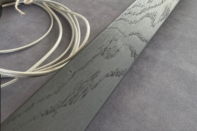 May include: Close-up of a dark gray wooden plank with a textured grain pattern. A silver-colored braided cord is coiled in the upper left corner. The wood has a distressed appearance, with dark streaks.