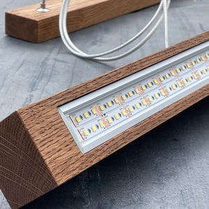 May include: A modern wooden light fixture with a rectangular frame. The fixture houses two rows of LED lights and is suspended by white cords. The wood has a natural grain and a warm brown color.