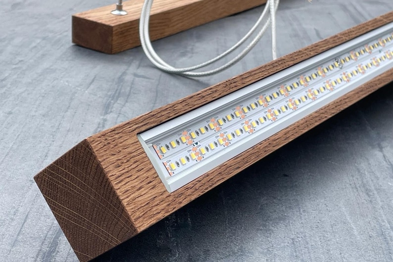 May include: A modern wooden light fixture with a rectangular frame. The fixture houses two rows of LED lights and is suspended by white cords. The wood has a natural grain and a warm brown color.