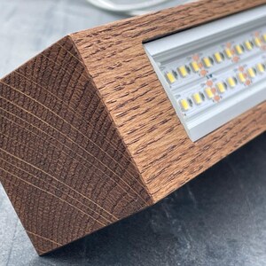 May include: A rectangular wooden light fixture with a warm brown finish. The fixture houses a series of bright LED lights, visible within a recessed metal channel. The wood grain is prominent, adding a natural aesthetic to the design.