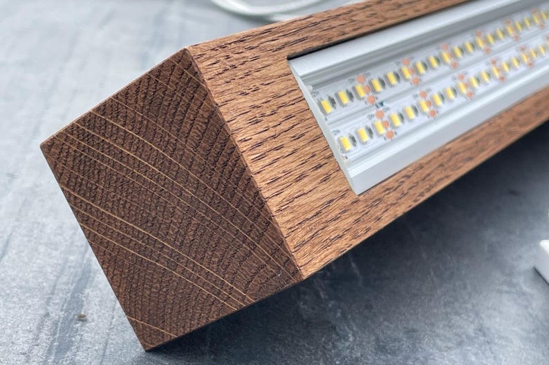 May include: A rectangular wooden light fixture with a warm brown finish. The fixture houses a series of bright LED lights, visible within a recessed metal channel. The wood grain is prominent, adding a natural aesthetic to the design.