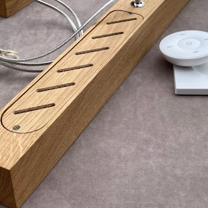May include: A rectangular wooden light fixture with a series of angled cutouts and a small metal switch. A white remote control and base are on the right. A silver cable is attached to the fixture.