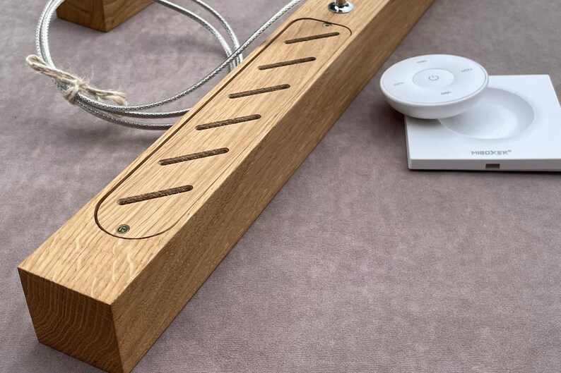 May include: A rectangular wooden light fixture with a series of angled cutouts and a small metal switch. A white remote control and base are on the right. A silver cable is attached to the fixture.