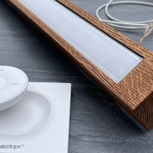 May include: A wooden LED light fixture with a white light panel and a white remote control. The fixture has a rectangular shape with a brown wood grain finish. The remote control has several buttons, including a power button. The brand name "MiBOXER" is visible.