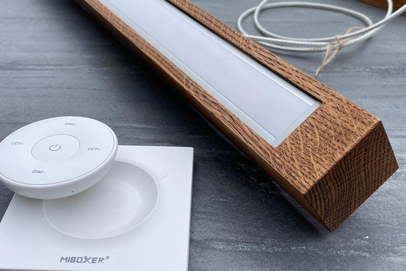 May include: A wooden LED light fixture with a white light panel and a white remote control. The fixture has a rectangular shape with a brown wood grain finish. The remote control has several buttons, including a power button. The brand name "MiBOXER" is visible.