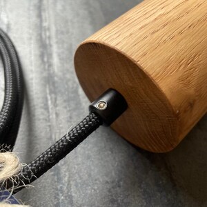 May include: Close-up of a cylindrical wooden pendant light fixture. The light has a natural wood finish and a black braided cord. The fixture is designed for indoor lighting.