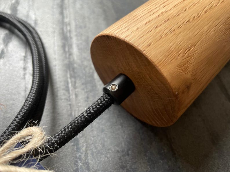 May include: Close-up of a cylindrical wooden pendant light fixture. The light has a natural wood finish and a black braided cord. The fixture is designed for indoor lighting.