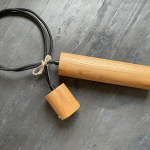 May include: A wooden pendant light fixture with a cylindrical shade and a smaller, conical socket. The light has a black cord and is tied with twine. The fixture is made of light-colored wood.