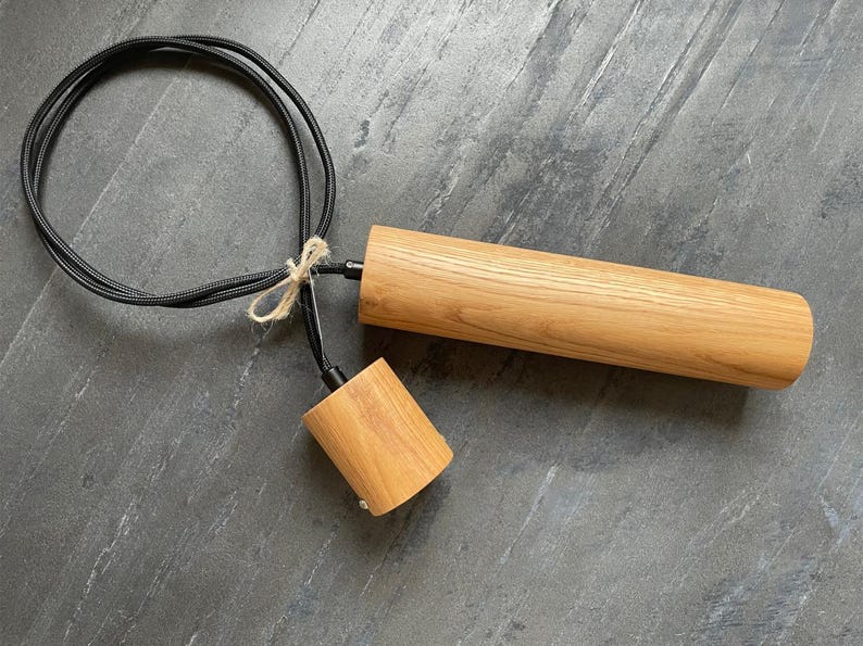 May include: A wooden pendant light fixture with a cylindrical shade and a smaller, conical socket. The light has a black cord and is tied with twine. The fixture is made of light-colored wood.