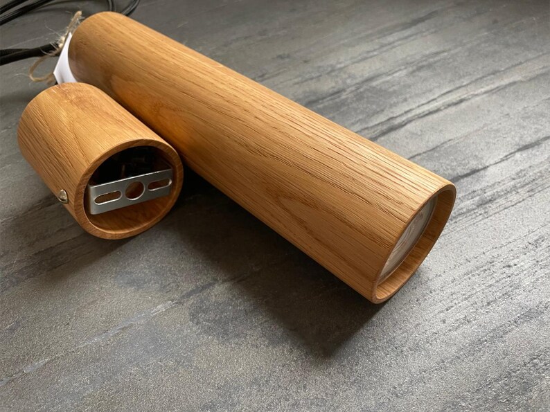 May include: A wooden cylindrical pendant light fixture with a natural wood grain finish. The fixture is composed of two parts: a long, cylindrical body and a smaller, circular base. The base has a metal mounting bracket. The light fixture is designed for ceiling installation.