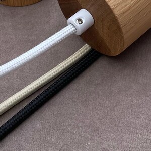May include: Close-up of a wooden cylindrical object with a white plastic connector. Three braided cords in white, beige, and black extend from the object. The cords are arranged diagonally against a neutral background.