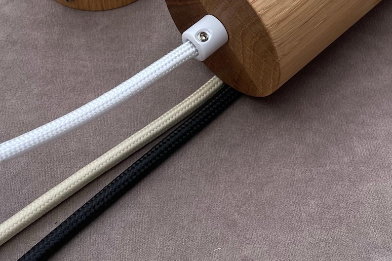 May include: Close-up of a wooden cylindrical object with a white plastic connector. Three braided cords in white, beige, and black extend from the object. The cords are arranged diagonally against a neutral background.