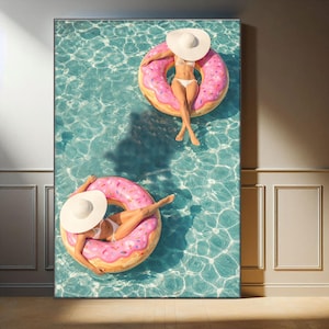 以下が含まれることがあります： プールでピンクのドーナツ型浮き輪に乗ってくつろぐ2人の姿を描いたキャンバスプリント。白い水着とつば広の帽子を着用。水は透明感のあるターコイズブルーで、夏のレジャーを連想させる。屋内展示。