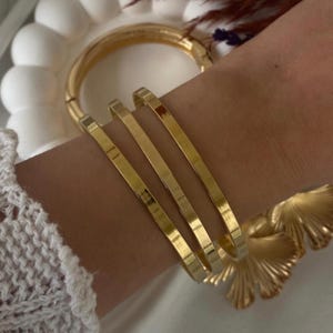 May include: Three gold bangles stacked on a wrist. The bangles are all different widths and have a smooth, polished finish.