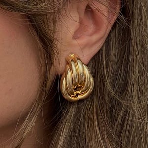 Può includere: Orecchini dorati e grossi con un design annodato. Gli orecchini sono di un colore oro brillante e lucido e hanno una forma arrotondata. L'immagine mostra una parte dell'orecchio e dei capelli.