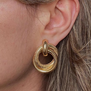 Peut inclure: Boucles d'oreilles créoles dorées à la finition texturée. Les boucles d'oreilles sont composées de plusieurs cercles entrelacés.