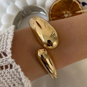 May include: A gold-colored cuff bracelet with a polished, reflective surface. The bracelet features two large, oval-shaped elements connected by a hinge. The design is minimalist and modern, suitable for various styles.