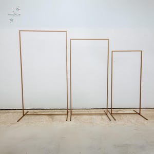 Może przedstawiać: Trzy złote metalowe stojaki łukowe w różnych rozmiarach. Stojaki wykonane są z cienkich metalowych rur i mają prosty, nowoczesny design.