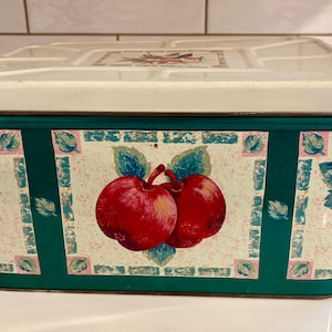May include: Rectangular metal storage tin with a cream-colored lid and green trim. The sides feature a repeating pattern of red apples, green leaves, and purple grapes. The lid has a floral design.