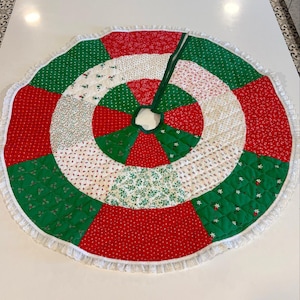 May include: A quilted Christmas tree skirt with a patchwork design. The skirt features red, green, and white fabric sections with floral patterns and a white lace trim. A green fabric tie is attached to the center opening.