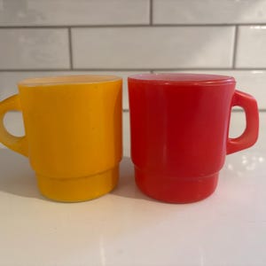 May include: Two stackable mugs made of smooth, opaque glass. One mug is a vibrant yellow with a rounded handle, while the other is a bright red with a squared handle. Both mugs feature a distinct stepped base design, creating a uniform silhouette.