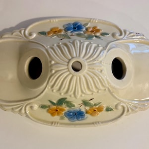 May include: Cream-colored ceramic ceiling light fixture with floral accents. The fixture has an oval shape with two circular openings and a central decorative element. The flowers are blue and yellow.