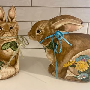 May include: Two decorative Easter bunny figures. One is upright, the other is seated. Both are brown with floral accents and ribbon details. The upright bunny has a green and cream ribbon, while the seated bunny has a blue ribbon.