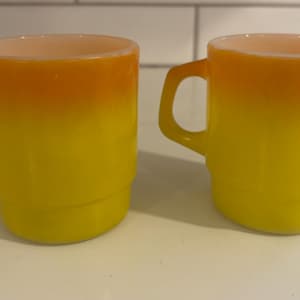 Puede incluir: Dos tazas vintage con un degradado de amarillo a naranja. Las tazas tienen un asa y una base apilada. El interior de las tazas es de color melocotón claro. Las tazas son de vidrio.