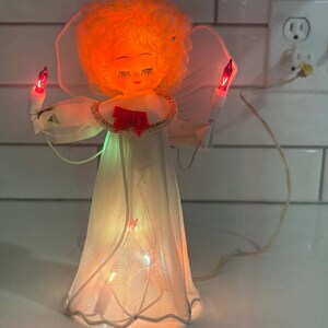 Puede incluir: Un adorno vintage para la parte superior del árbol de Navidad con forma de ángel. El ángel tiene un vestido blanco, un halo y sostiene dos luces de vela rojas. La cara del ángel está iluminada con un brillo cálido, y el vestido está iluminado desde dentro.