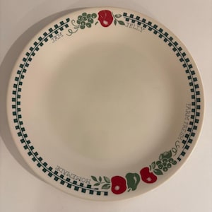 Puede incluir: Plato de cerámica blanquecina con un borde a cuadros verdes y negros. El plato presenta ilustraciones de manzanas rojas y uvas verdes, con las palabras "JAM JELLY" y "FARM FRESH & CO. HOMEMADE" impresas en el borde.