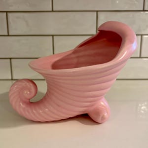 Può includere: Un vaso a forma di cornucopia in ceramica rosa. Il vaso ha un design a spirale e poggia su tre piedini. L'apertura è ampia e curva. Il vaso è di colore rosa pastello.